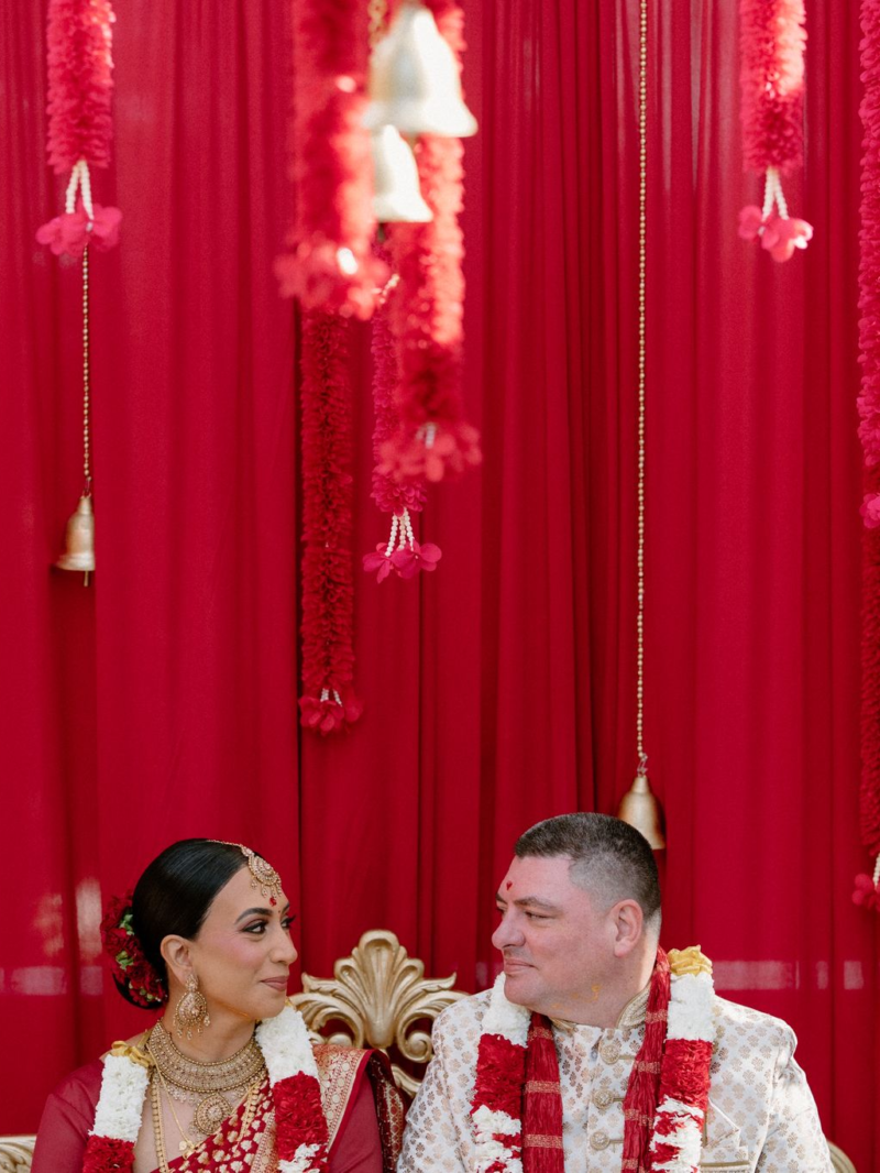 Anushka and Dan seated before red drapery at their South Indian Hindu wedding ceremony at Springfield House, Dural