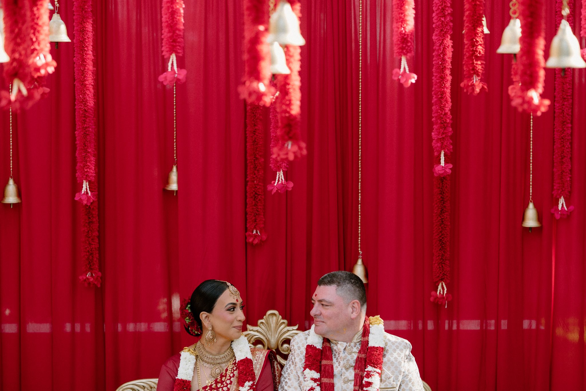Anushka and Dan seated before red drapery at their South Indian Hindu wedding ceremony at Springfield House, Dural