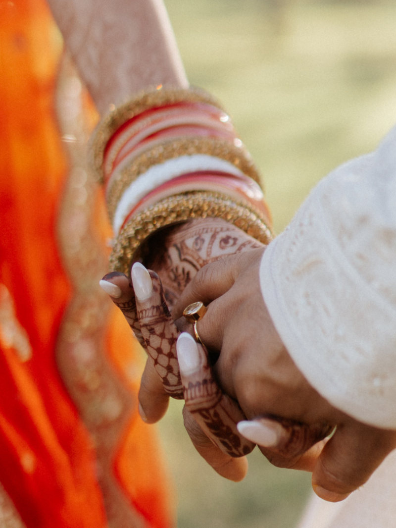 A bride and groom hold hands, her mehndi-covered fingers stacked with red, white and gold bangles.