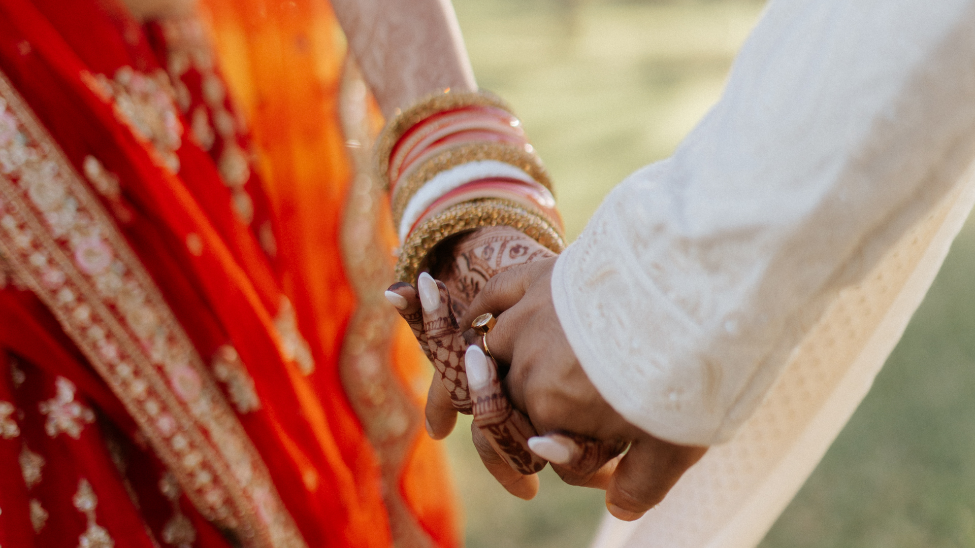 A bride and groom hold hands, her mehndi-covered fingers stacked with red, white and gold bangles.